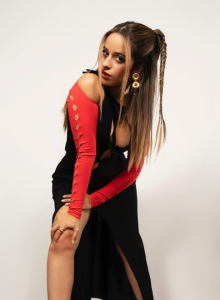 Cantante Camila Cabello posa frente a un fondo blanco con un vestido negro y una camiseta roja por dentro y en su cabeza lleva dos coletas medias con trenzas
