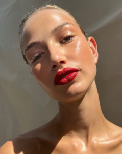 Closeup de modelo con cabello recogido, maquillaje glowy y labios rojos usando eyelighter en el parpado