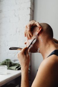 hombre de espaldas se rasura con cuchilla y espuma de afeitar frente a una ventana en un baño