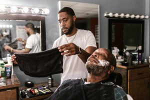 En una barbería un barbero extiende una toalla junto a un hombre recostado en una silla esperando a ser rasurado