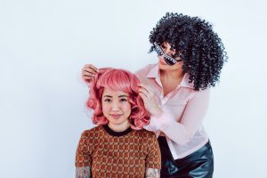 una chica con peluca de color negro rizada y blusa rosa le coloca a otra chica una peluca de color rosa