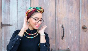 chica con gafas aparece sonriendo con un pañuelo en la cabeza de colores, collares de colores y una blusa de cuello color negro sobre un fondo de madera